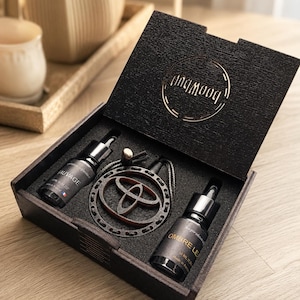 Könnte beinhalten: Eine schwarze Holzgeschenkbox mit zwei kleinen Glastropfflaschen mit den Aufschriften Sauvage und Ombre Leather sowie einem runden Holzanhänger mit einem mittigen Ausschnitt, alles in einer schwarzen Schaumstoffeinlage.