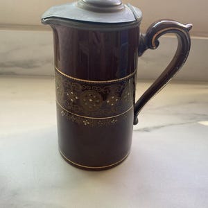May include: A dark burgundy ceramic pitcher with a lid and handle. The pitcher features gold-colored decorative bands with floral patterns. The lid is a darker metal with a small decorative figure on top. The handle has gold accents.
