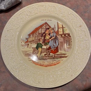 Puede incluir: Un plato de cerámica color crema con un borde floral en relieve. El centro presenta una ilustración colorida titulada CRIES OF LONDON, que muestra a tres figuras cerca de un carro de madera. El texto inferior dice ROUND AND SOUND FIVEPENCE A POUND DUKE CHERRIES.