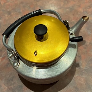 May include: A silver-toned metal kettle featuring a bright gold lid with a black knob. The kettle has a black handle, a small black lever on the side, and a spout. A small circular stamp with the text Made in Japan is visible on the metal base.