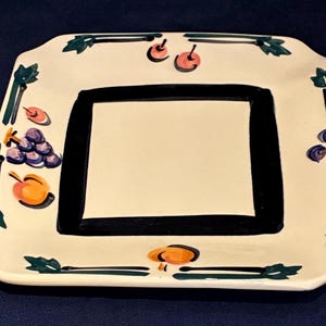 May include: A square ceramic plate with a cream-colored base featuring a thick black square border in the center. The rim is decorated with hand-painted motifs of purple grapes, orange peaches, pink cherries, and green leaves arranged along the edges.