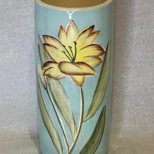 May include: A cylindrical ceramic vase with a light blue glazed finish. The exterior features a hand-painted illustration of a large yellow lily with brown-tipped petals and long green leaves extending from the base towards the top rim.