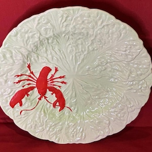 Puede incluir: Una bandeja de servicio redonda de cerámica con una superficie moldeada y texturizada que imita la apariencia de una hoja de col verde. Una langosta de color rojo brillante está grabada en el lado izquierdo del plato, contrastando con el diseño de follaje verde pálido.