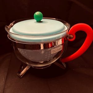 Könnte beinhalten: Eine Teekanne aus klarem Glas mit einem hellblauen Deckel und einem grünen, kugelförmigen Knauf. Die Kanne hat einen leuchtend roten Griff, ein Metallband um die Mitte und drei Metallfüße. Ein weißes Innensieb ist sichtbar und das Wort bodum ist an der Seite aufgedruckt.