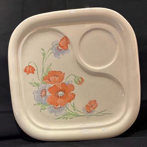 May include: A square off-white ceramic snack plate with rounded corners, featuring a recessed circular section for a cup. The surface is decorated with a floral pattern of orange and light purple poppies with green stems and leaves.