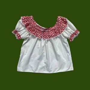 Peut inclure: Chemise blanche de style paysan avec un imprim&eacute; floral rouge et blanc autour de l'encolure. L'encolure est fronc&eacute;e et orn&eacute;e d'une dentelle blanche.