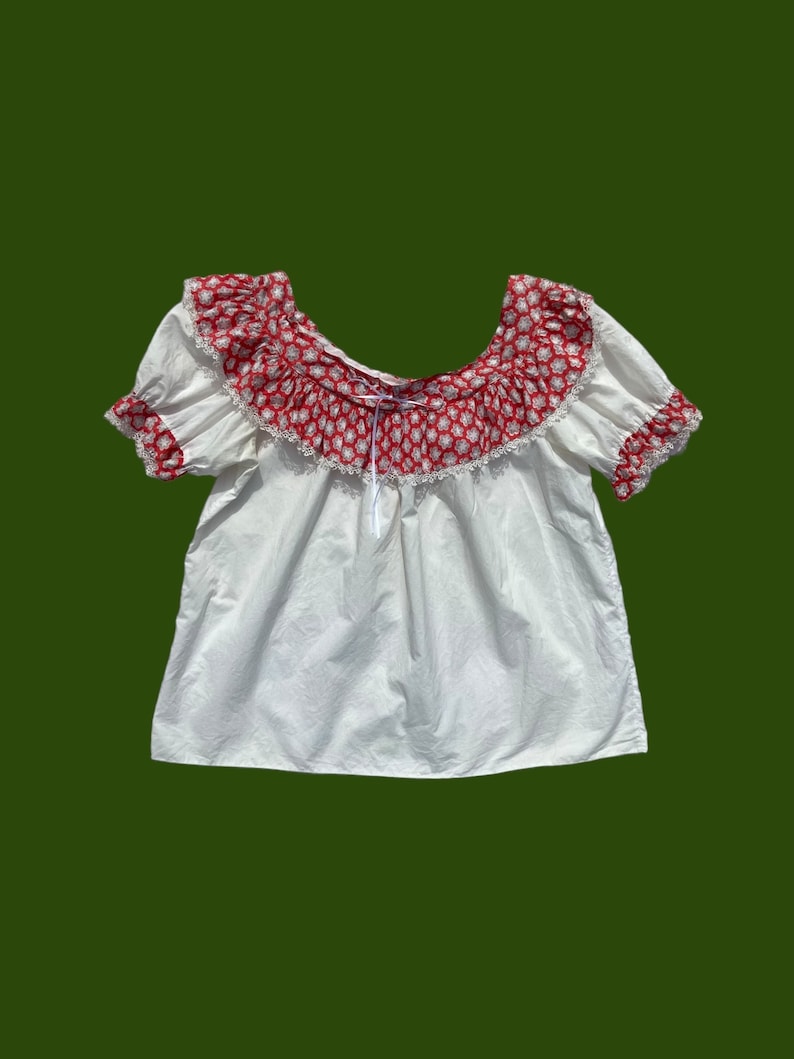 Peut inclure: Chemise blanche de style paysan avec un imprim&eacute; floral rouge et blanc autour de l'encolure. L'encolure est fronc&eacute;e et orn&eacute;e d'une dentelle blanche.
