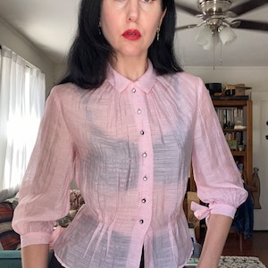 Puede incluir: Una blusa de botones de color rosa claro transparente con cuello fruncido y mangas largas. La blusa tiene una cintura ajustada y un dobladillo ligeramente acampanado. Las mangas tienen un detalle de lazo en los puños.