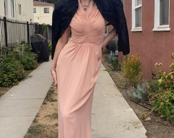 May include: A long, blush-colored dress with a pleated bodice and a flowing skirt. A black jacket is draped over the shoulders. The dress is paired with metallic shoes. The overall style is vintage-inspired.