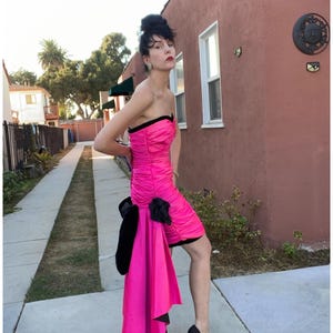 May include: A hot pink strapless dress with a black bow and a black train. The dress is made of a shiny fabric and has a fitted bodice and a flowing skirt.