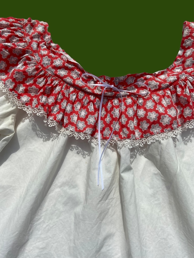 Peut inclure: Chemise blanche avec un col &agrave; volants imprim&eacute; floral rouge et blanc et une bordure en dentelle blanche. Le col est dot&eacute; d'un ruban blanc.