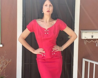May include: A vintage, bright red two-piece dress. The top has a scoop neckline and short sleeves, with pearl-like buttons. The pencil skirt is knee-length. The dress is worn by a person standing in front of a brown door.