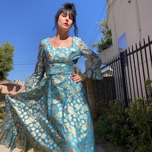 May include: A long, turquoise dress with gold floral patterns. The dress features long sleeves with flared cuffs, a square neckline, and a fitted waist. The skirt flows to the ground. The dress is made of a shiny fabric.