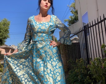 May include: A long, turquoise dress with gold floral patterns. The dress has long sleeves with flared cuffs, a square neckline, and a fitted waist. The skirt flows to the ground. The dress is made of a shiny fabric.