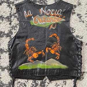 Könnte beinhalten: Schwarze Lederweste mit handgemaltem Skorpion- und Landschaftsdesign. Die Weste trägt den Text "La Novia Queretaro" in Orange und Weiß. Seitliche Schnürung und ein klassischer Biker-Stil.