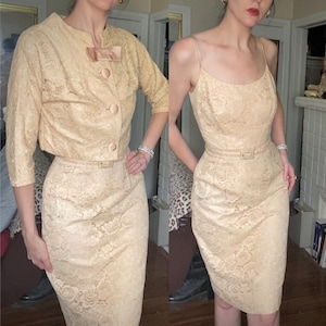 May include: A two-piece dress set in a cream colour with a lace pattern. The dress has a fitted bodice and a skirt that falls to the knees. The jacket has a short, cropped style and three buttons down the front.