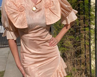 May include: A vintage peach-colored dress with a unique design. The dress features a ruffled skirt, a ruched bodice, and a cape-like top with puffed sleeves and decorative rosettes. The dress is made of a shiny, silky material.