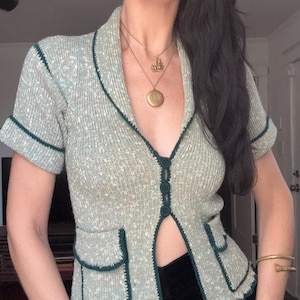 May include: A short-sleeved knit cardigan in a speckled light green and white yarn. The garment features a deep V-neckline, a dark green trim along the edges, three central buttons, and two front pockets with dark green flap detailing.