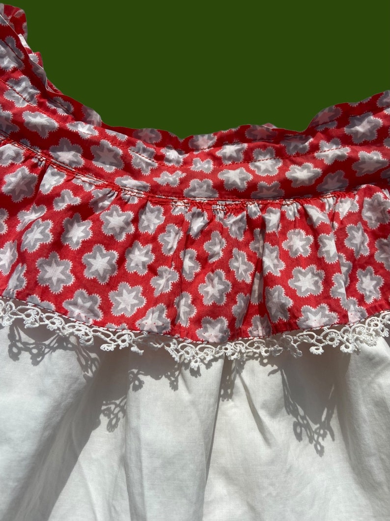 Peut inclure: Une robe en coton blanc avec un volant &agrave; l'encolure imprim&eacute; de fleurs rouges et blanches. Le volant est bord&eacute; de dentelle blanche.