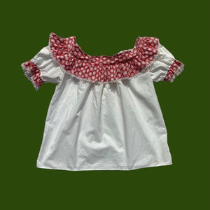 Peut inclure: Haut blanc de style paysan avec un imprim&eacute; floral rouge et blanc autour de l'encolure. Le haut a des manches courtes et un corsage pliss&eacute;.