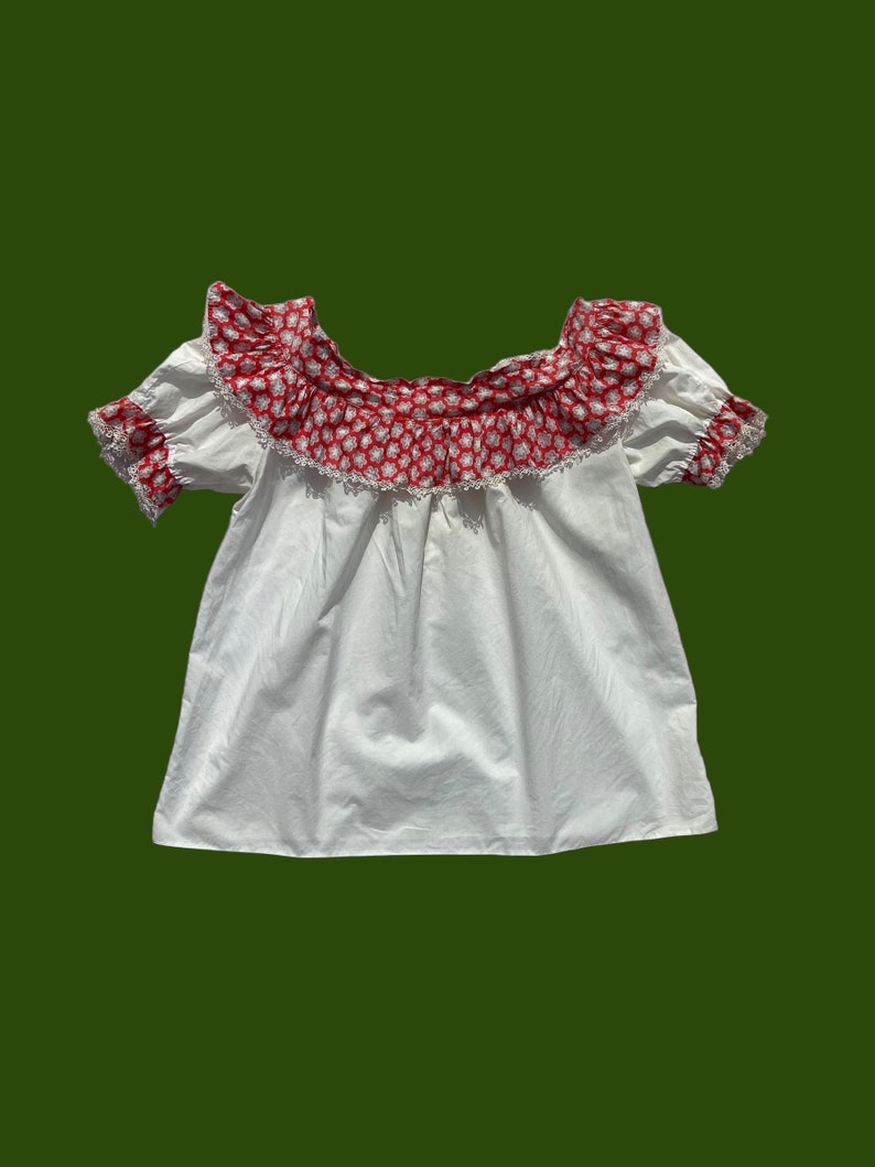 Peut inclure: Haut blanc de style paysan avec un imprim&eacute; floral rouge et blanc autour de l'encolure. Le haut a des manches courtes et un corsage pliss&eacute;.
