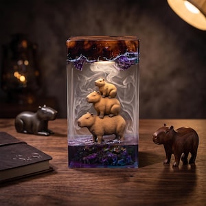 May include: A rectangular resin lamp featuring three stacked capybara figurines inside a clear block. The top and bottom sections contain dark, swirling purple and brown resin textures. A soft internal light illuminates the capybaras and smoke.