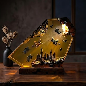 May include: A trapezoidal resin lamp with a warm yellow glow, featuring a miniature castle, figures on broomsticks, and flying ghosts embedded inside. The piece is mounted on a dark wooden base, creating a scene of a magical school.