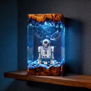 Op de afbeelding: Een rechthoekige lamp met een centrale R2-D2-droidfiguur ingesloten in heldere hars. De boven- en onderkant zijn gemaakt van natuurlijk hout met een wervelende nerf. Het interieur gloeit met blauw licht en verlicht sliertige, rookachtige patronen.