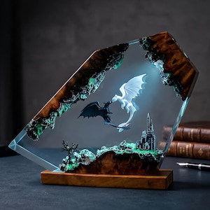 May include: A decorative resin and wood sculpture featuring a black dragon and a white dragon facing each other. The scene includes a miniature castle and a bare tree set within a clear resin frame with glowing green accents and a wooden base.