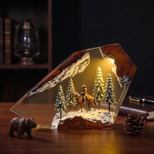Puede incluir: Una lámpara decorativa de resina y madera con una escena en miniatura de una persona montando a caballo en un bosque de pinos nevado. La pieza está iluminada desde el interior con una luz cálida que resalta los bordes de madera y la base.
