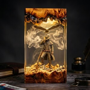 Puede incluir: Una lámpara rectangular de resina con base y parte superior de madera, que encierra una escena en miniatura de un vaquero con abrigo largo y rifle, sobre terreno rocoso. Un águila calva aparece en vuelo sobre la figura, rodeada de humo.