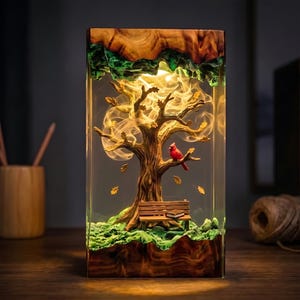 May include: A rectangular resin lamp featuring a natural wood base and top, enclosing a detailed miniature scene of a tree with a red cardinal perched on a branch, a wooden bench, a small book, and floating leaves, illuminated by warm internal lighting.