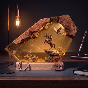 May include: A decorative resin and wood sculpture featuring a miniature cowboy on a rearing horse atop a rocky outcrop, with a flying eagle, desert plants, and wisps of smoke inside the amber-toned resin, set on a rectangular wooden base.