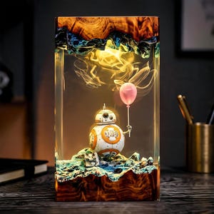 Op de afbeelding: Een rechthoekige lamp van hars en hout met een witte en oranje BB-8 droid die een roze ballon vasthoudt. De droid staat op een getextureerde blauwe en groene basis in een blok heldere hars, verlicht door een warm schijnend licht.