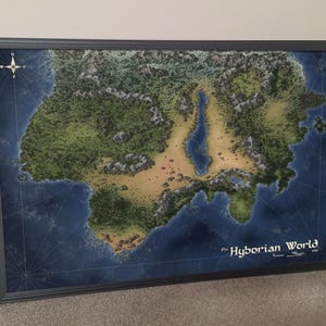 May include: Framed map of the Hyborian World, featuring a detailed topographical depiction of a fictional landmass. The map is framed in a dark gray border, with a compass rose in the upper left corner. The title "Hyborian World" is written in white.