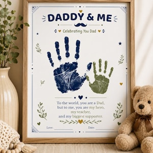 May include: A framed print titled DADDY & ME featuring a large navy blue adult handprint and a smaller olive green child handprint. The text reads: To the world, you are a Dad, but to me, you are my hero, my teacher, and my biggest supporter.