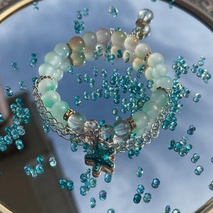 May include: A beaded bracelet with light blue and white beads, accented with silver chain links and a teal starfish charm. The bracelet is displayed on a reflective surface scattered with blue crystals, creating a shimmering effect. The bracelet is a piece of jewelry.