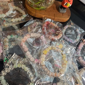 May include: A collection of beaded bracelets in individual clear plastic packaging, featuring various pastel-colored translucent beads, metallic spacers, and small decorative charms including a Christmas tree, a shooting star, and a palm tree.