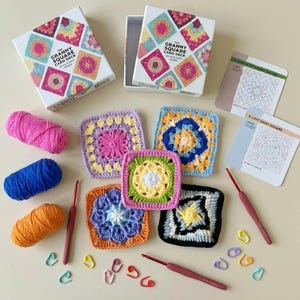May include: A Granny Square Card Deck box set with instructional cards, five crocheted squares in various colors, three skeins of yarn in pink, blue, and orange, two crochet hooks, and several colorful plastic stitch markers for crafting.