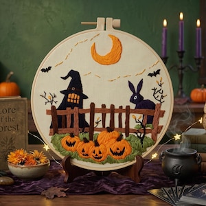 May include: A circular embroidery hoop art piece featuring a Halloween scene with a crescent moon, a witch hat, a black rabbit, a wooden fence, and carved jack-o'-lanterns on a cream fabric background, displayed on a wooden stand.