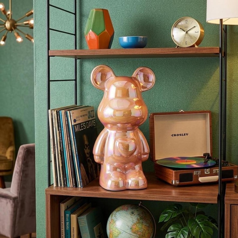 May include: A pearlescent pink bear figurine with a heart on its chest sits on a wooden shelf. Other items on the shelf include a vintage record player, a gold clock, and a stack of vinyl records. A green and orange vase and a blue bowl are also visible.