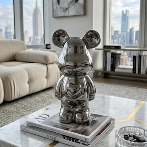 May include: A shiny silver bear figurine with a heart-shaped detail on its chest. The figurine is displayed on a marble coffee table, atop a stack of magazines. The background features a cityscape view through a large window.