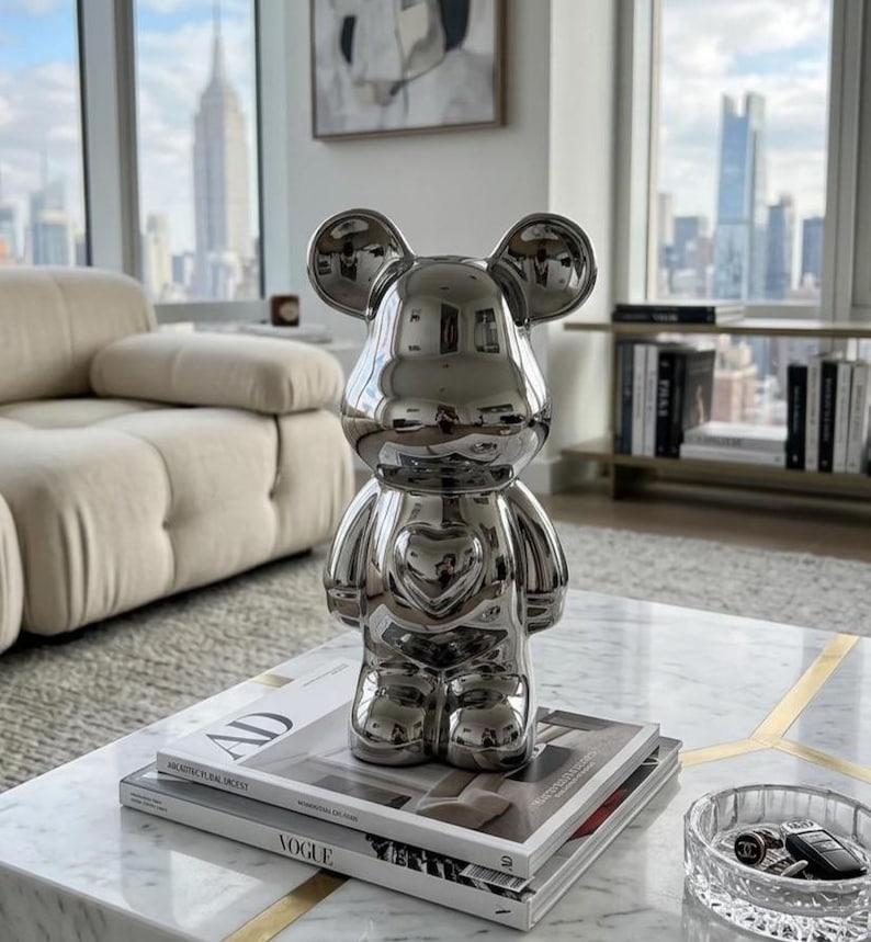 May include: A shiny silver bear figurine with a heart-shaped detail on its chest. The figurine is displayed on a marble coffee table, atop a stack of magazines. The background features a cityscape view through a large window.