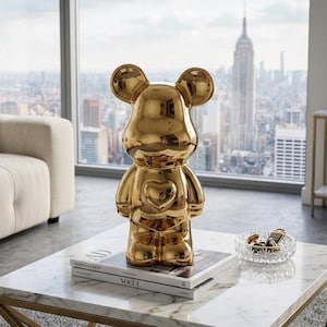 May include: A gold-colored bear-shaped figurine with a heart-shaped detail on its chest. The figurine is displayed on a white marble coffee table, with a stack of books and a small glass dish. The background features a cityscape.