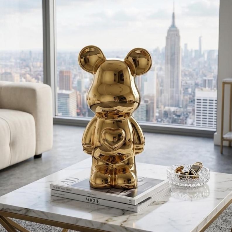 May include: A gold-colored bear-shaped figurine with a heart-shaped detail on its chest. The figurine is displayed on a white marble coffee table, with a stack of books and a small glass dish. The background features a cityscape.