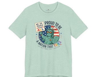 May include: A light green heathered t-shirt featuring a graphic of the Statue of Liberty wearing rainbow sunglasses. The text reads: WE THE PEOPLE MEANS ALL, PROUD TO BE A NATION THAT TRIES, LIBERTY JUSTICE EQUALITY DIGNITY FOR ALL, LOVE > HATE.