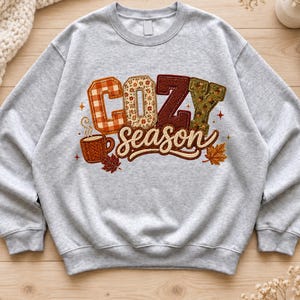 May include: A light grey crewneck sweatshirt featuring a graphic print with the text COZY season. The letters are filled with various autumn-themed patterns including orange plaid, floral, red herringbone, and green acorn prints, with a coffee mug.