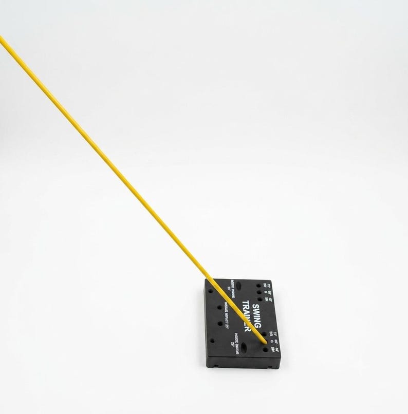 May include: A black rectangular swing trainer base with white text and multiple circular holes, featuring a long, thin, bright yellow rod extending diagonally upward from one of the holes. The text on the base reads SWING TRAINER.