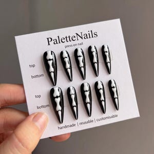 May include: A set of ten black press-on nails with a white skeleton finger bone design arranged on a white display card labeled PaletteNails. The card includes text indicating top and bottom orientation, handmade, reusable, and customisable.