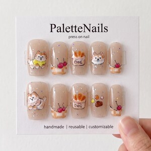 Può includere: Un set di dieci unghie finte color beige con finitura glitterata, caratterizzate da illustrazioni dipinte a mano di cani, ciliegie e cestini. La scheda di presentazione riporta PaletteNails e il testo handmade, reusable, customizable.